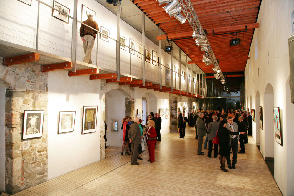 Einblick bei der Eröffnung der Ausstellung in der Burg von Ljubljana, an den Wänden hängen Bilder in der Mitte des Raumes stehen Menschen, die diese betrachten und sich unterhalten.